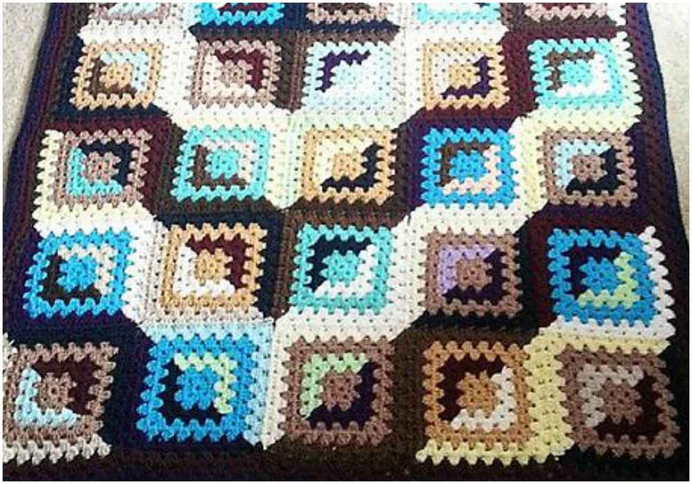 A vibrant crochet blanket featuring a geometric granny square pattern in shades of blue, brown, cream, and gray.