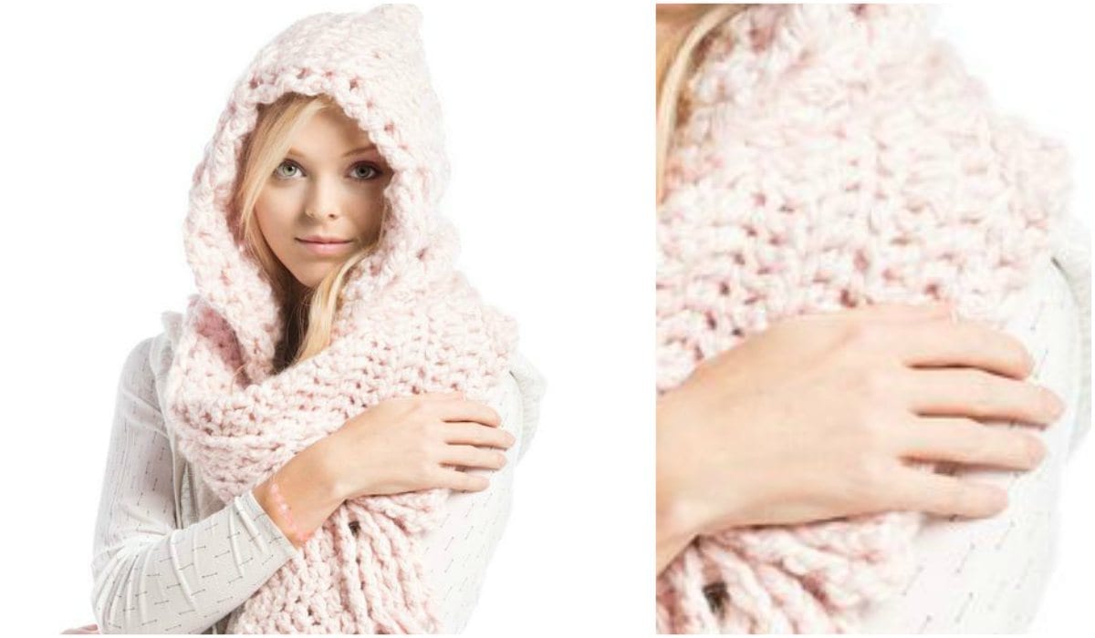 A person wearing a large, knitted pink hooded scarf with a chunky texture holds it gracefully in one hand, showcasing its intricate crochet pattern.