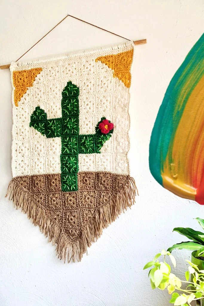 Crocheted wall hanging of a green cactus with a red flower, brown base, yellow corners, and fringe at the bottom, hanging on a white wall next to a colorful abstract paintingโperfect as crochet decorations for fall.