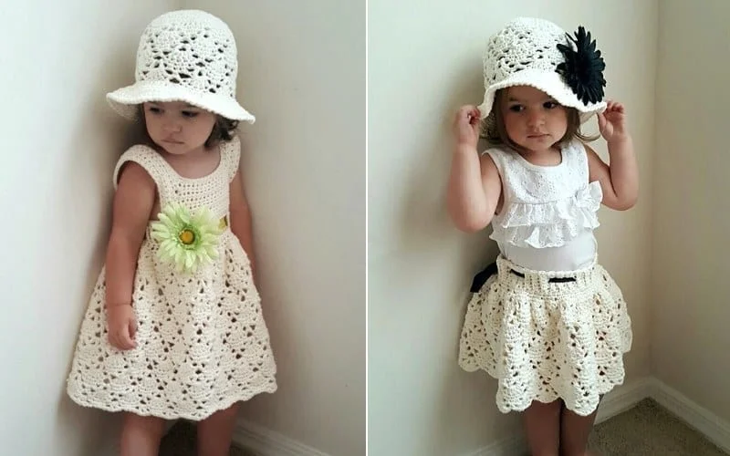 Two toddlers in charming white crochet outfits from Little Lady Clothes, one adorned with a green flower on the dress and the other with a black flower on the hat, stand against a pristine white wall, showcasing their timeless style.