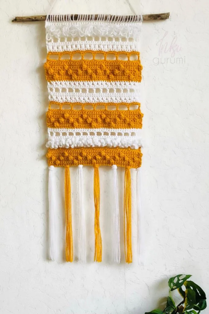 A handmade wall hanging with alternating white and yellow crocheted stripes, attached to a wooden stick, with long yarn tassels hanging from the bottomโperfect as crochet decorations for fall.