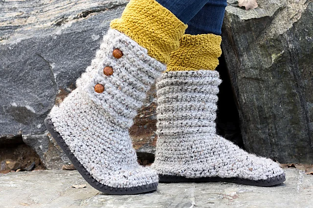 Person wearing chunky crocheted gray Cabin Boots with Flip Flop Soles, accented with brown buttons and yellow knit socks, standing outdoors on a stone surface.