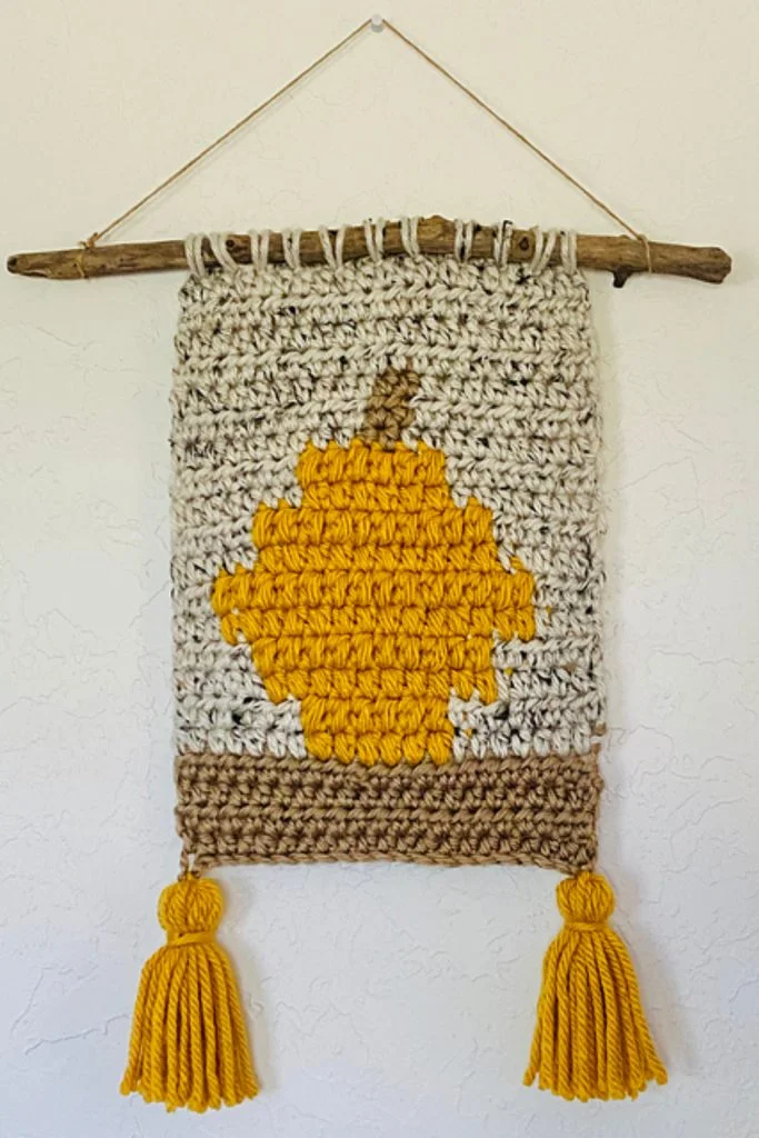 A crocheted wall hanging with a yellow pumpkin design, hung on a stick and decorated with two yellow tassels at the bottomโperfect as crochet decorations for fall to add a cozy seasonal touch to your space.