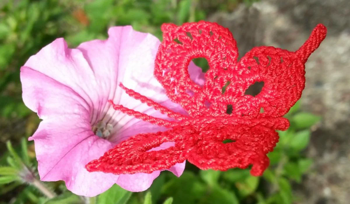 A pink petunia flower with a red, carefully crocheted butterfly perched beside it, surrounded by lush green foliage, captures a serene moment.