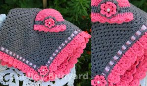 Gray and pink crocheted baby blanket and hat, adorned with delicate flower decorations, rest beautifully on a white chair surrounded by lush plants. The cozy set features a charming granny square crochet pattern, adding a touch of timeless elegance.