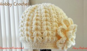 A cream-colored crocheted hat featuring a textured puff stitch pattern and a large flower embellishment on the side, reminiscent of a beanie hat design, displayed indoors.