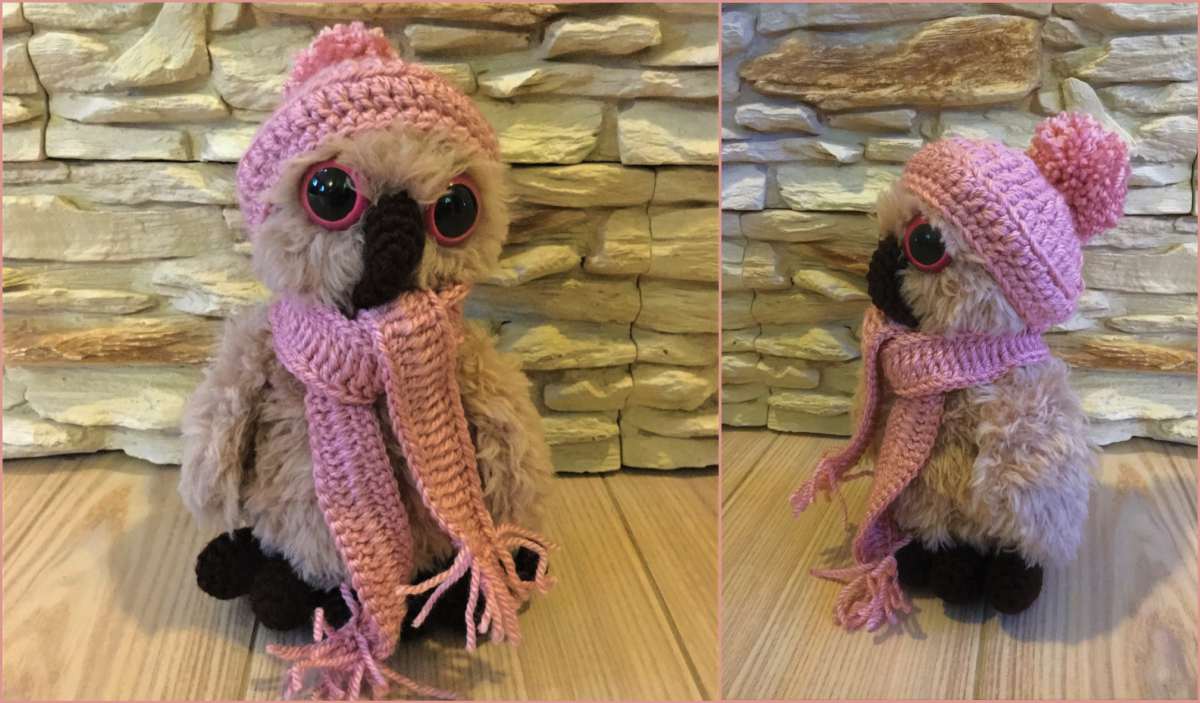 A plush, wintery owl dons a pink crocheted hat and scarf, perched gracefully on a wooden floor against a textured stone wall.