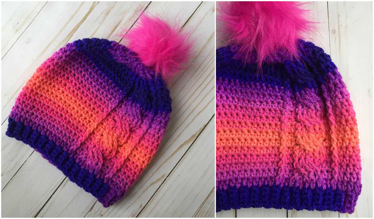 A Rora Cable Slouch beanie in a gradient of purple, pink, and orange hues, topped with a bright pink pom-pom, is displayed on a light wooden surface. This XL adult crochet pattern is perfect for adding a pop of color to your winter wardrobe.