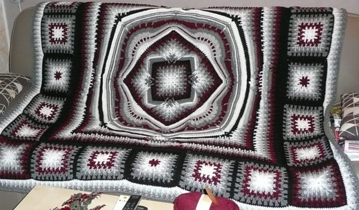 A crocheted blanket with a geometric pattern in red, white, and black is elegantly displayed on a sofa, complemented by matching CAL Crochet Square pillows.