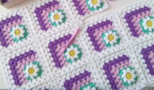 Crochet blanket adorned with delightful daisy granny squares in shades of purple, pink, green, and white. A pink crochet hook rests on the blanket. Perfect for those who love following a cozy crochet tutorial to create something beautiful and unique.