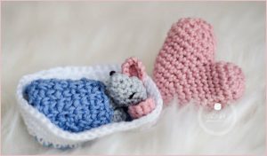 A small crocheted gray mouse, crafted with an amigurumi crochet pattern, sleeps under a blue blanket beside a pink heart on a white fluffy surface.