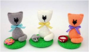 Three charming cat amigurumi sit on green bases: a gray cat with a mouse, a white one with a yarn ball, and an orange cat beside a red bowl labeled "cat." This delightful souvenir set inspires creativity and charm in every detail.