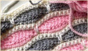 A close-up of a crocheted pattern using pink, gray, and white yarn showcases the wave-like elegance of the Millstone Stitch.