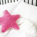 A pink, crocheted Sirius Star Pillow rests on a neatly made bed with white sheets and pillows.