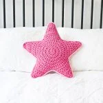 A pink crocheted Sirius Star Pillow is placed on a made bed with white bedding and a slatted headboard.