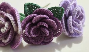 Three crocheted roses in shades of purple with green leaves displayed on a light background invite creativity. Discover the artistry with a free pattern to guide your own crochet roses creation.