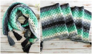 Introducing the Stormy Day Wrap: a crochet scarf with green, gray, and black stripes, accented by sleek black tassels. Artfully displayed on a wooden surface, this piece combines comfort and style. Perfect as an accessory or crafted through a free crochet pattern for personal flair.