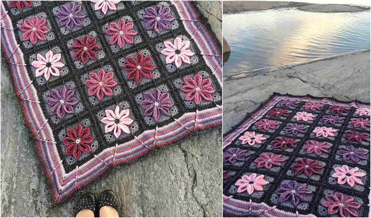 A crocheted blanket with flower motifs in shades of pink and purple is displayed on a rocky surface near water, accompanied by an adorable set of matching baby booties. Black shoes are visible in the foreground.