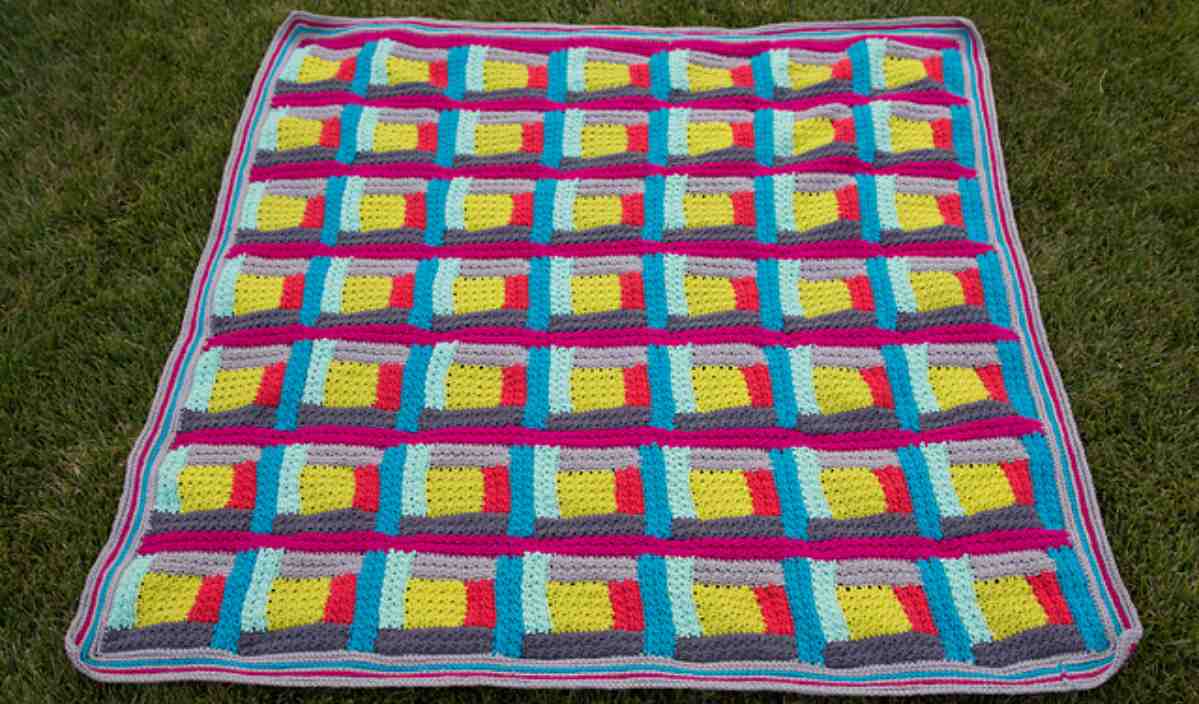 A colorful Log Cabin Blanket with a geometric pattern featuring rectangles in blue, yellow, red, and gray, spread out on green grass.