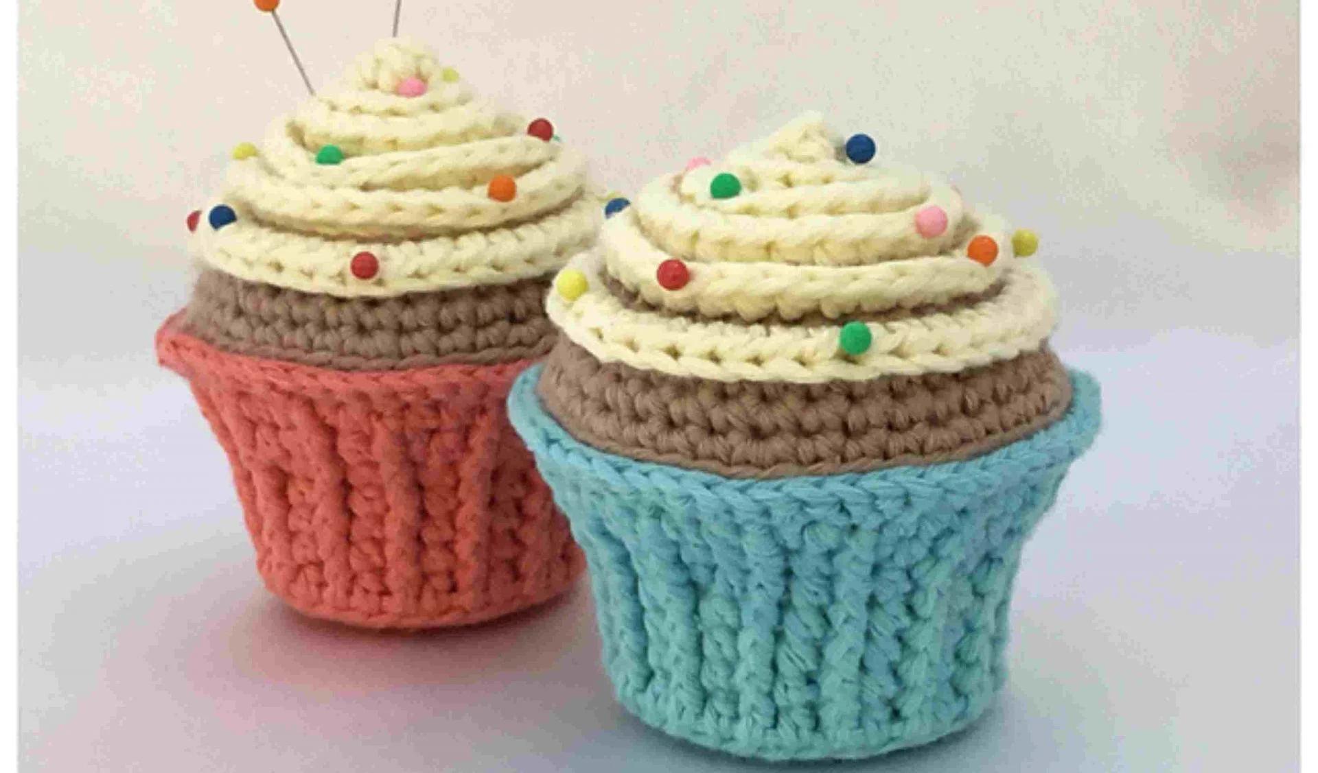 Two delightful crocheted cupcakes, one in a blue wrapper and the other in a red, serve as whimsical pin cushions adorned with colorful sprinkles and pins. Explore our free crochet pattern to craft your own charming cupcake pincushion set!