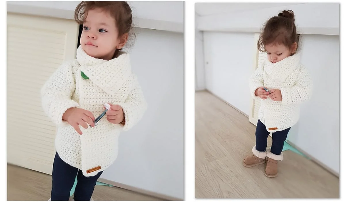 A toddler wearing a Snowdrop Cardigan, dark pants, and boots stands indoors holding an object.