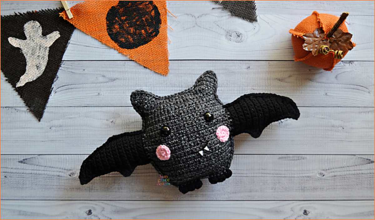 A crocheted bat kawaii with rosy cheeks and fangs sits on a wooden surface, surrounded by Halloween-themed decorations. This adorable cuddler is perfect for adding a whimsical touch to your spooky setup. Get creative and make your own with the free crochet pattern!