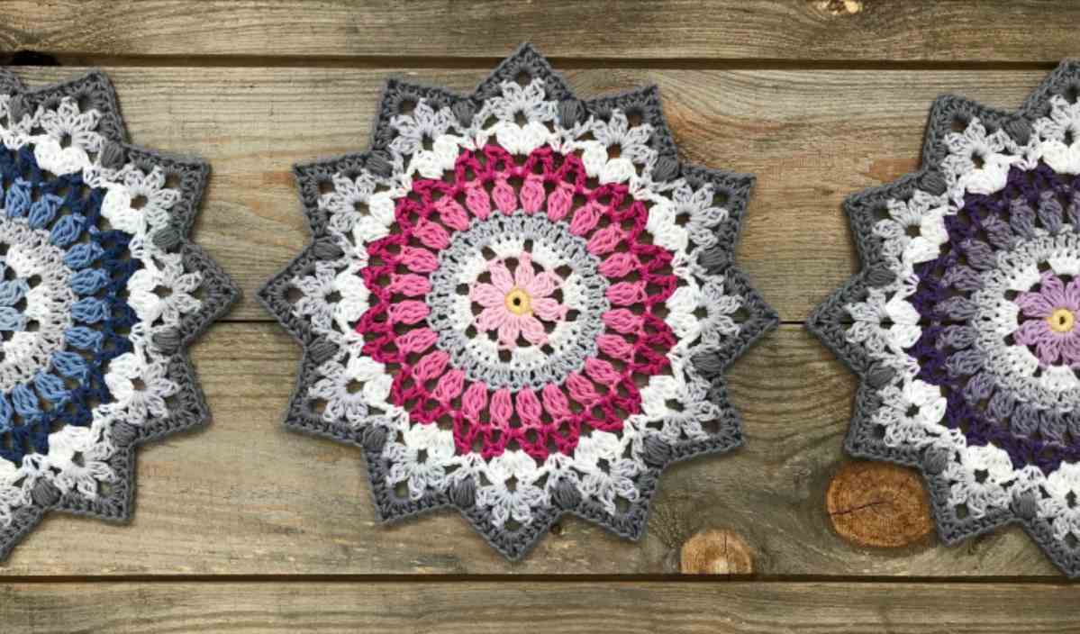 Three winter mandalas in blue, pink, and purple hues are displayed on a wooden surface, showcasing a beautiful crochet pattern.