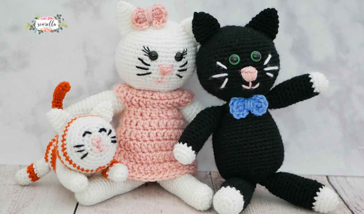 Three crocheted cat dolls: one white with a pink dress and bow, one black with a blue bow tie, and one small orange-striped cat. Each doll is as charming as an Ana Lucia Shawl, crafted with love from a delightful free crochet pattern.