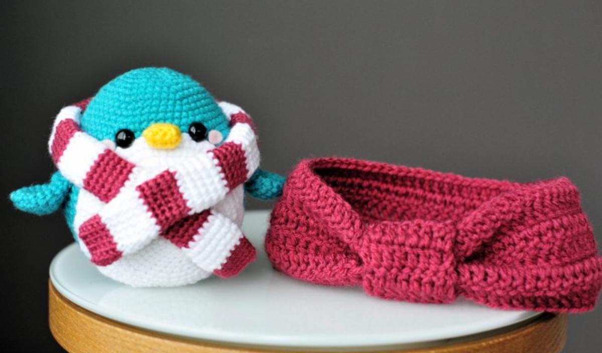 A crocheted penguin with a red and white scarf sits next to a red crocheted headband on a round wooden table, alongside an in full bloom design from the free crochet pattern collection.