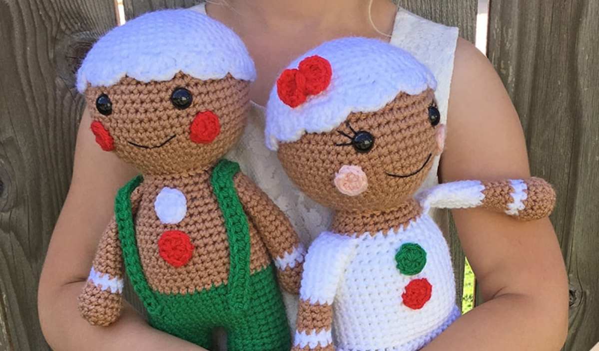 A person holds two crochet gingerbread dolls, one wearing green overalls and the other in a white outfit with a red flower. These delightful gingerbread people make for an adorable craft. If you're inspired, consider searching for a free crochet pattern to create your own festive friends!