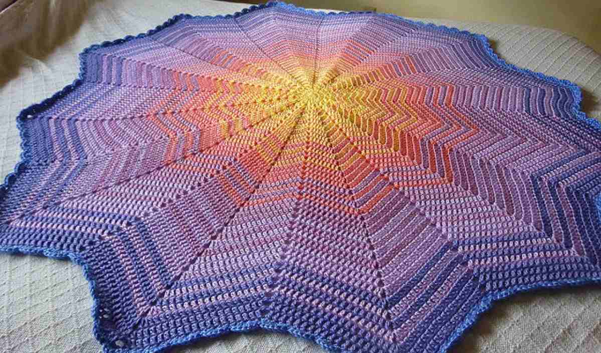 A star-shaped crochet blanket, reminiscent of a Sunrise Baby Ripple Afghan, displays a captivating gradient from yellow to purple. Laid flat on the surface, this piece beautifully showcases its intricate crochet pattern.