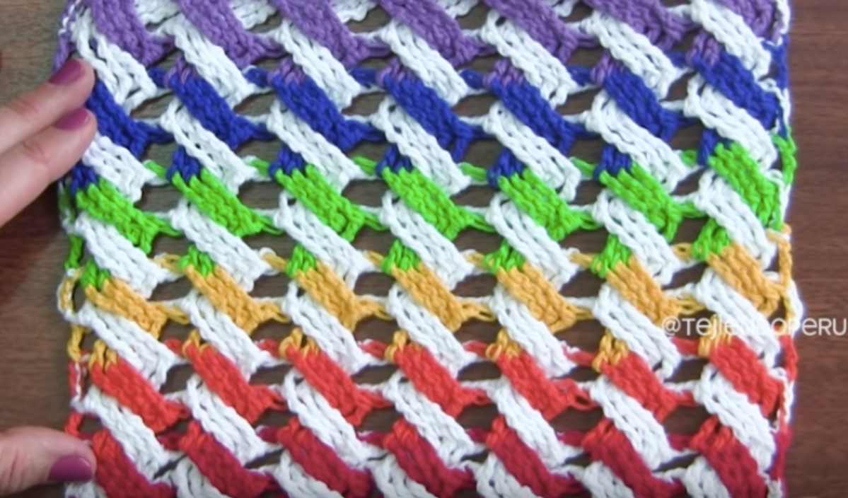 A vibrant C2C scarf showcasing a colorful woven knit pattern with diagonal stripes in purple, blue, green, yellow, and red on a white background, held by a hand on the left edge.