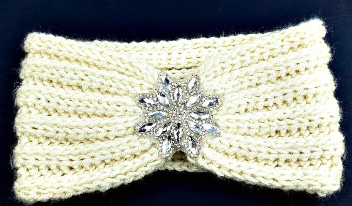 A cream-colored knitted headband with a textured crochet pattern and a silver, leaf-shaped jeweled brooch in the center.