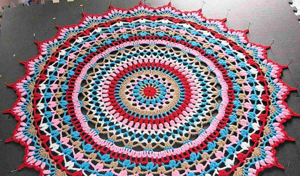 A colorful circular mandala from the Summer Splendor Crochet Pattern, featuring intricate designs in red, pink, blue, and tan, laid flat and pinned at the edges against a dark backdrop.