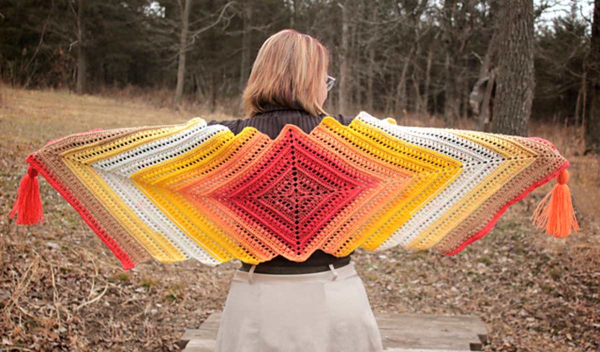 A person stands in a forest wearing a colorful, crochet shawl with shades of red, orange, yellow, and white, spread out like wings. Inspired by an easy crochet pattern, this vibrant accessory feels as playful as a cute penguin waddle.