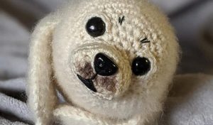 A crocheted white seal plush with large black eyes and a small black nose rests on a soft, gray surface, reminiscent of a Victorian lattice square's intricate elegance.
