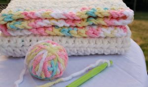A crocheted blanket with multicolored yarn sits on a table beside a crochet hook, a ball of matching yarn, and a crochet pattern that inspires creativity.