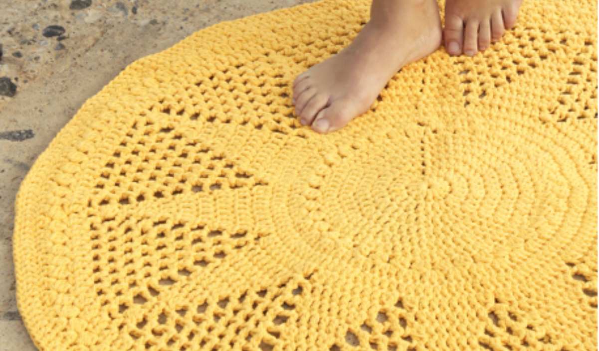 Sunny Rug: This yellow crochet rug with a radiant sunburst pattern rests on a sandy surface, perfectly embodying the spirit of a bright day. A person's feet are partially visible, adding warmth to this charming sunny scene.