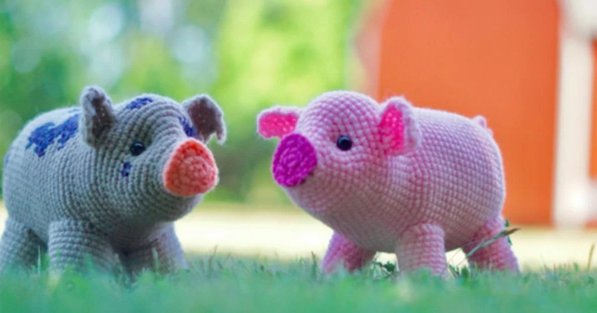 Two adorable crochet pigs, one gray with blue spots and the other pink, stand on grass with an orange structure in the background.