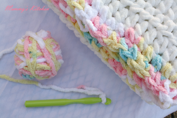 A crochet hook, a ball of pastel-colored yarn, and a partially crocheted blanket with chunky stitches are laid out on a white surface, perfect for making an Easy Beginner's Baby Blanket.