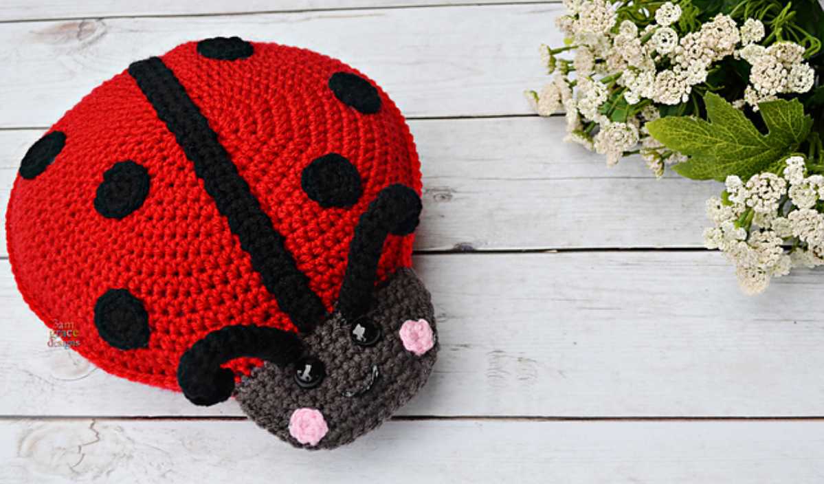 A crocheted ladybug pillow with a red body, black spots, and a smiling face rests on a wooden surface next to a cluster of white flowers. For those inspired by this creation, explore our free crochet pattern for matching accessories.