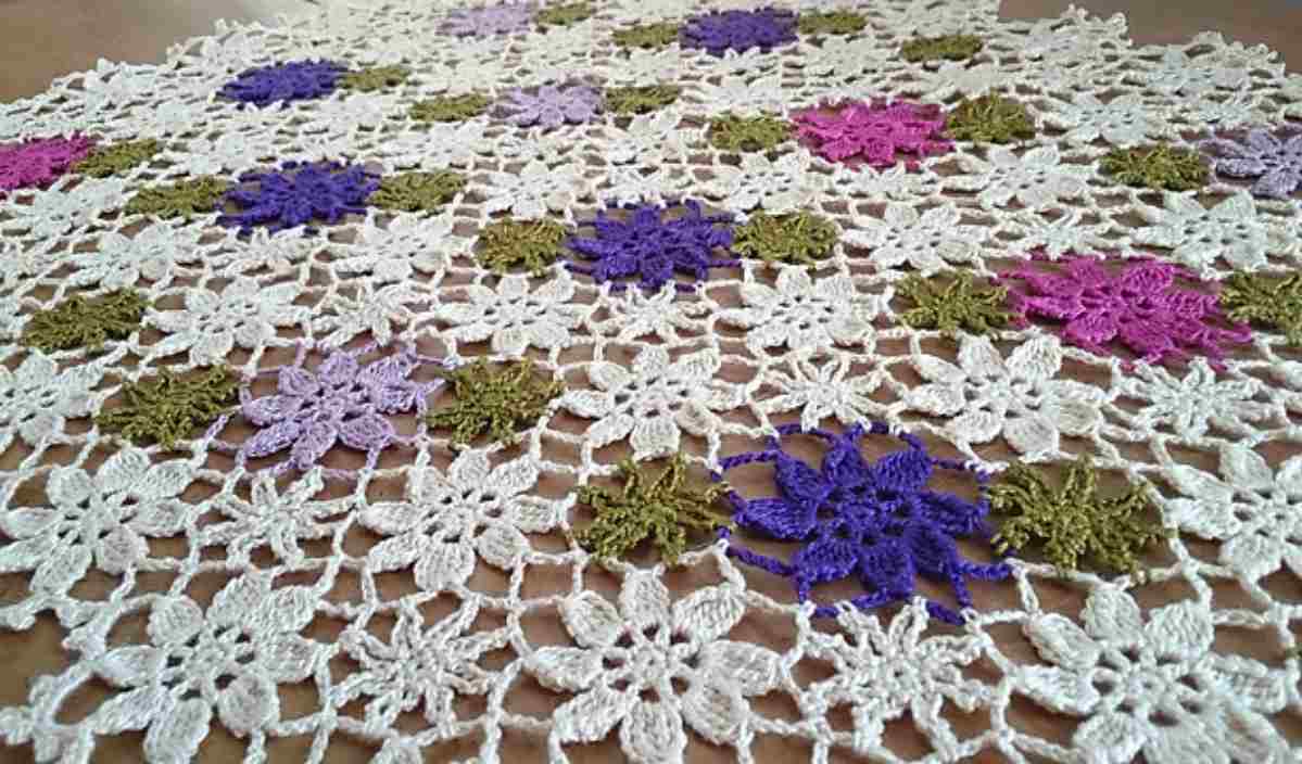 Crocheted lace fabric with a floral mandala design in white, purple, green, and pink on a flat surface.