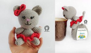 A crocheted gray cat with a red bow and skirt, elegantly sits on a hand, balancing on a yarn ball beside a brown coffee cup. This delightful scene is inspired by a Persian Dream crochet pattern.