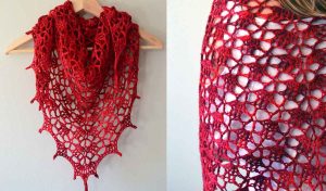 A vibrant red crocheted shawl with a lace pattern is displayed hanging on a hanger and draped over a person's shoulder, featuring an elegant flow reminiscent of an asymmetrical hem tunic.
