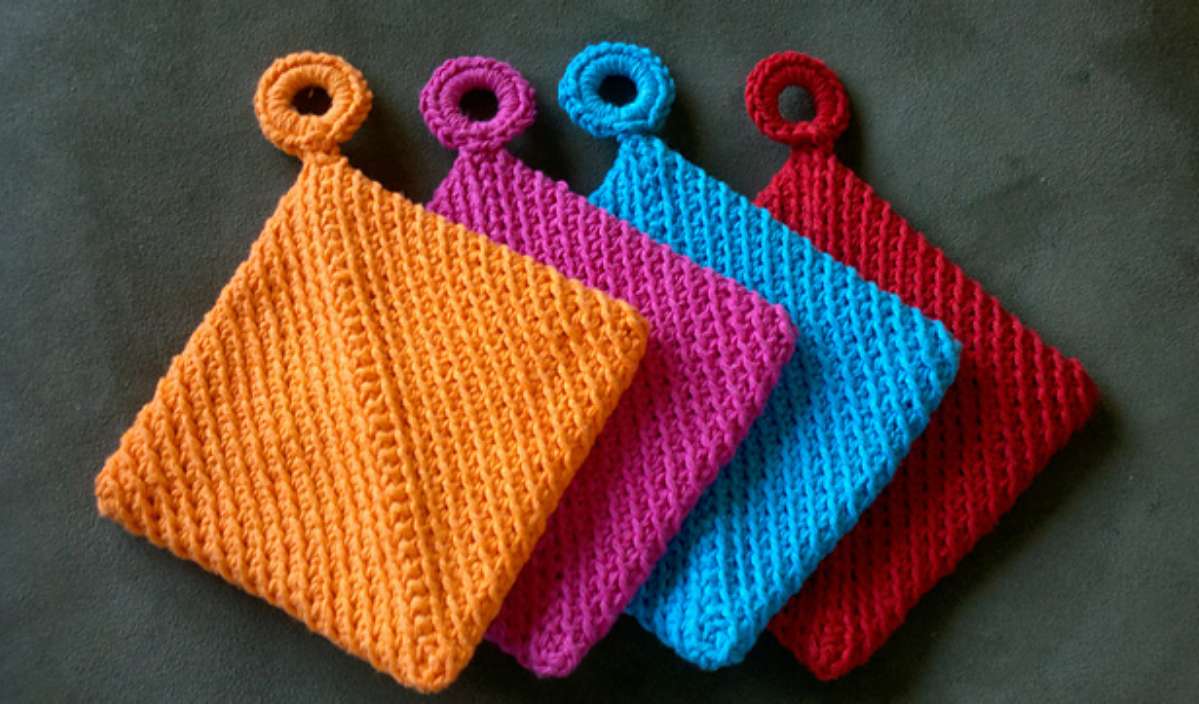 Four knitted pot holders in vibrant hues of orange, pink, blue, and red, each with a loop for hanging, are laid out in a row. Discover the rich texture reminiscent of a folded potholder that adds charm to your kitchen accessories.