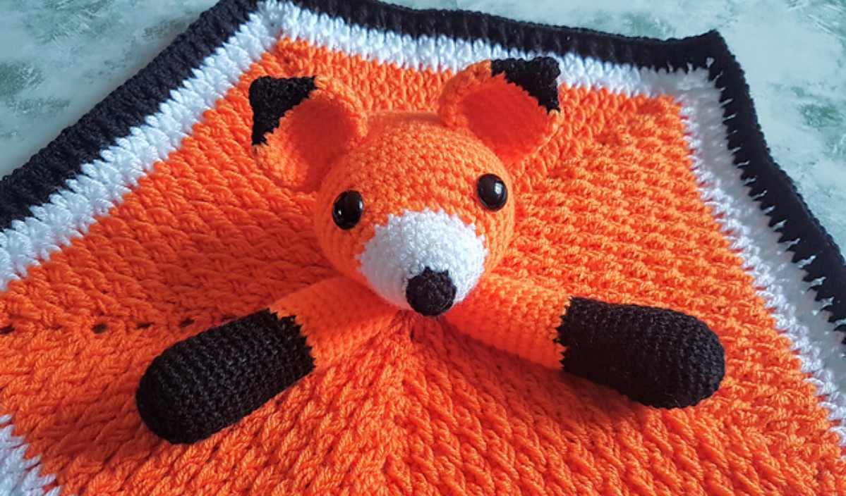 A crochet blanket showcasing an orange and black fox design with a 3D fox head and limbs, laid out on a flat surface. This charming piece is available as a free pattern for those wanting to craft their own unique fox security blanket.