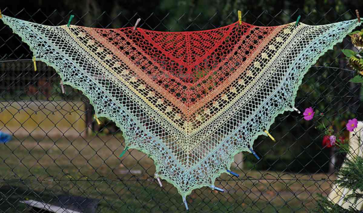 A colorful knitted shawl with red, orange, yellow, and green hues hangs on a fence beside a display of round crochet coasters.