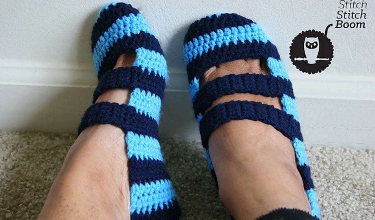 Feet wearing double strapped crochet slippers with alternating blue and navy stripes, against a white wall and beige carpet. Logo with an owl and the text "Stitch Stitch Boom" in the corner.