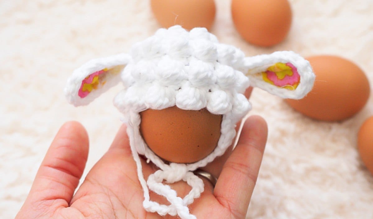 A hand holds a brown egg sporting a crocheted white hat with ears, surrounded by more eggs, reminiscent of charming sunflower crochet patterns.