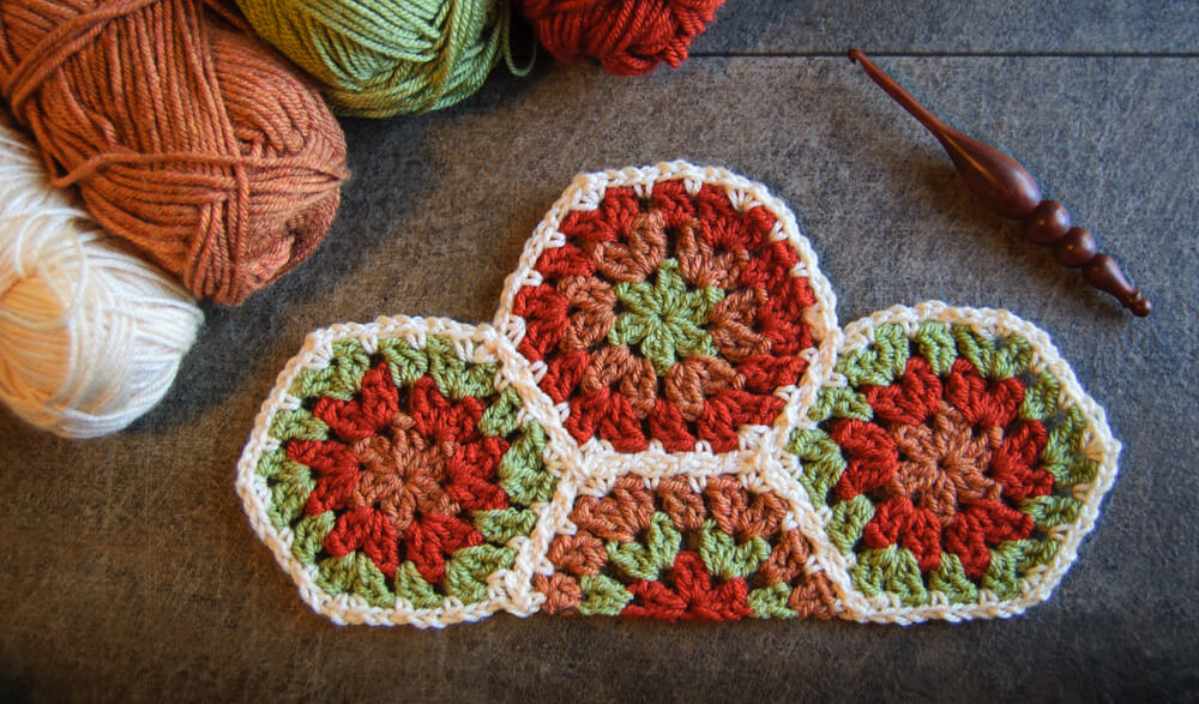 Three colorful crocheted hexagons connected together, bordered by balls of yarn in orange, brown, green, and beige, with a wooden crochet hook resting nearby on a gray surface. Explore free crochet patterns to create your own vibrant rainbow appliqué designs.